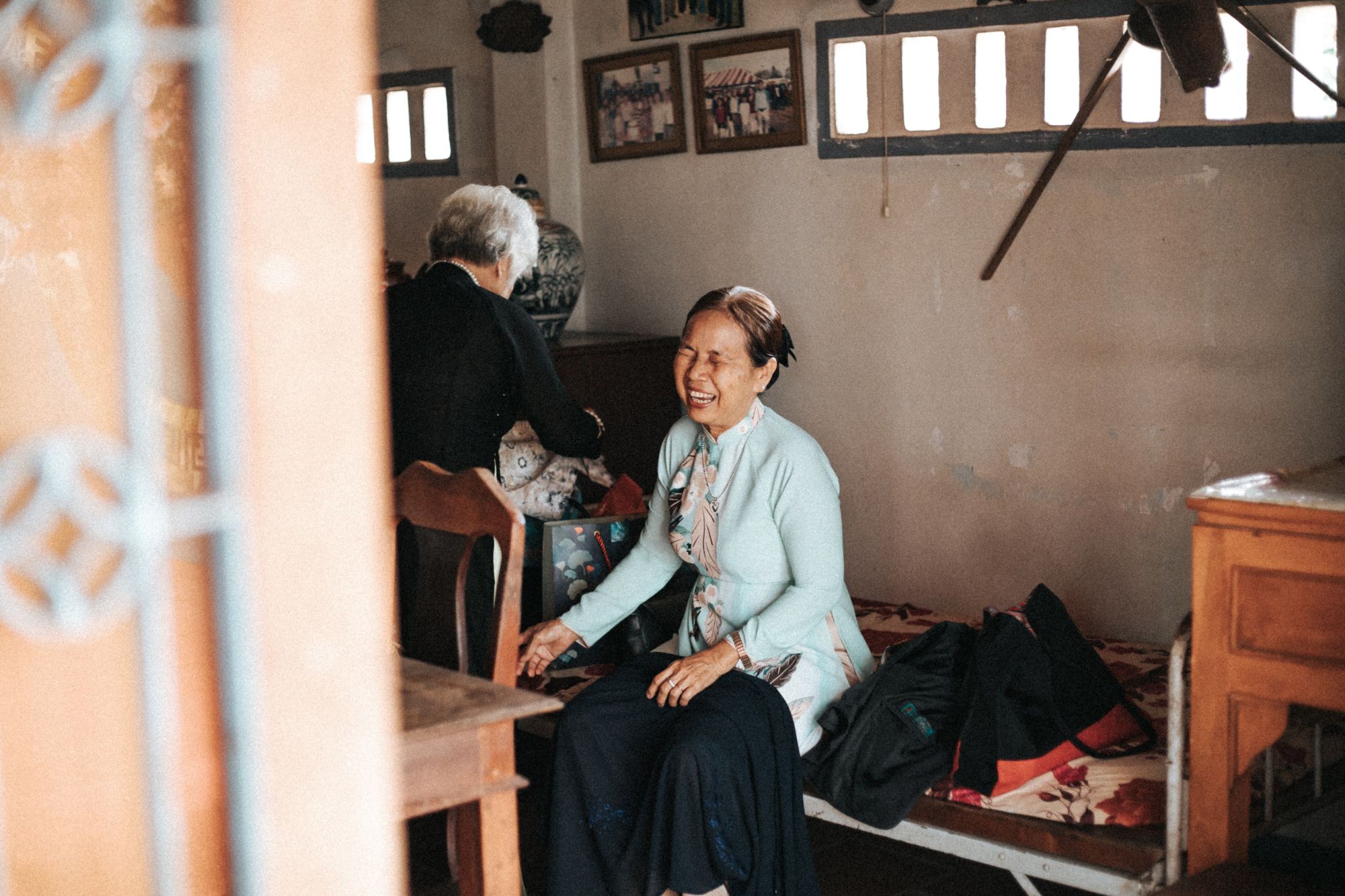 A woman laughing at home, seated on a daybed, while a friend looks through a bag