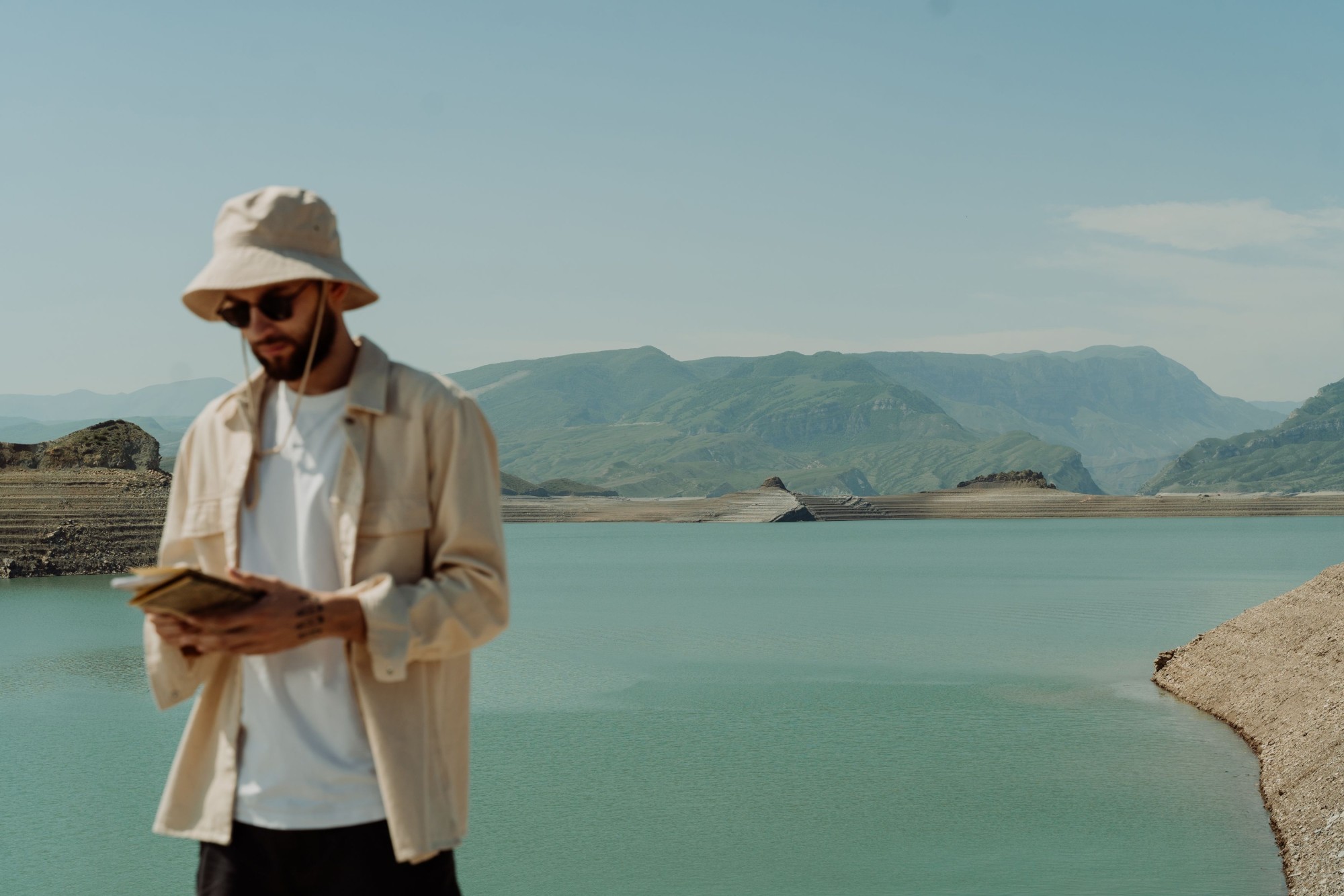 A traveler in a bucket hat reading a guidebook by a mountain lake